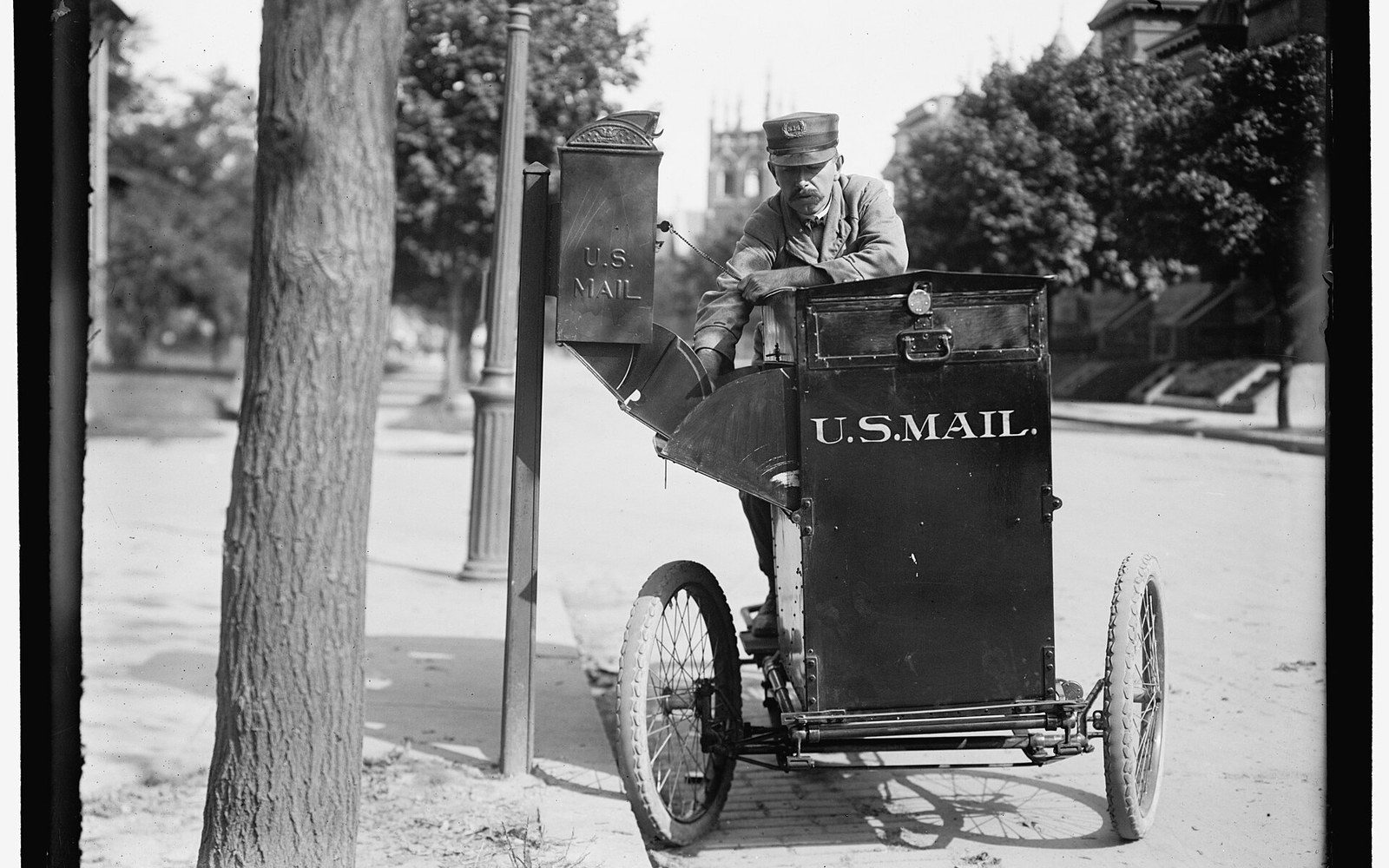 A uniformed postal worker empties a curbside mailbox into a small three-wheeled U.S. Mail cart