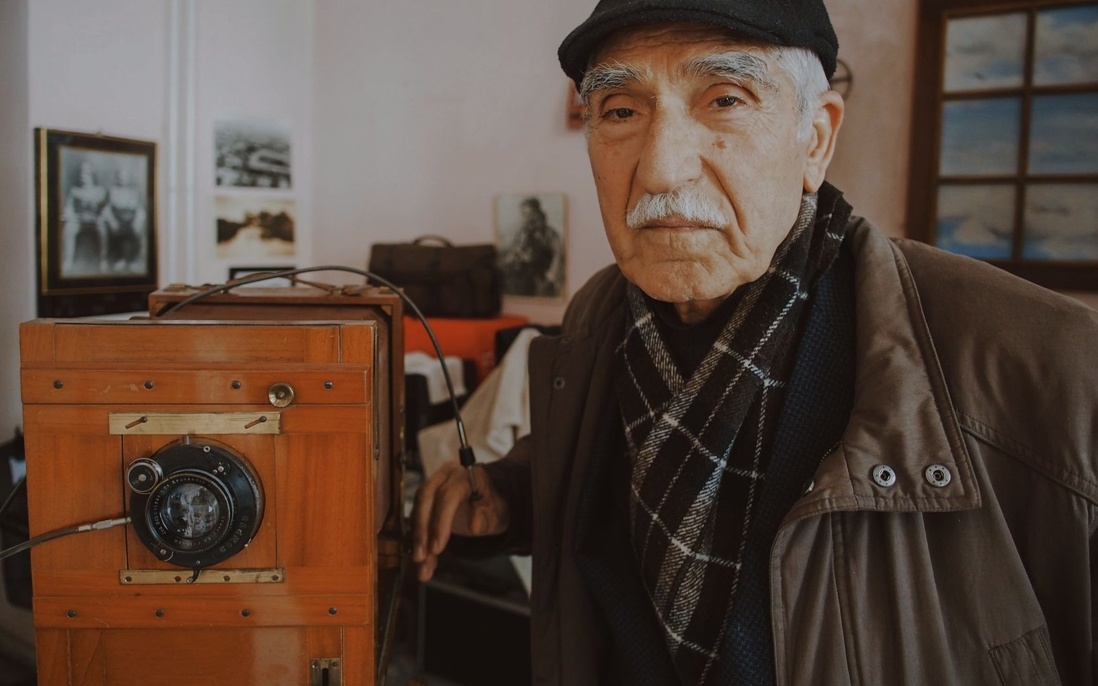 An elderly man in a flat cap stands beside a vintage wooden box camera